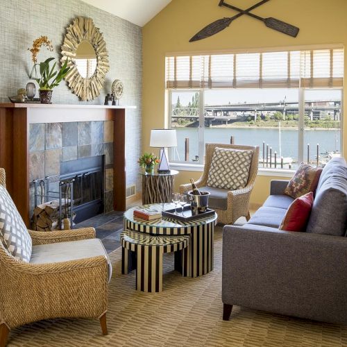 A cozy living room with cushioned seating, wicker chairs, a fireplace, and a large window overlooking a river.