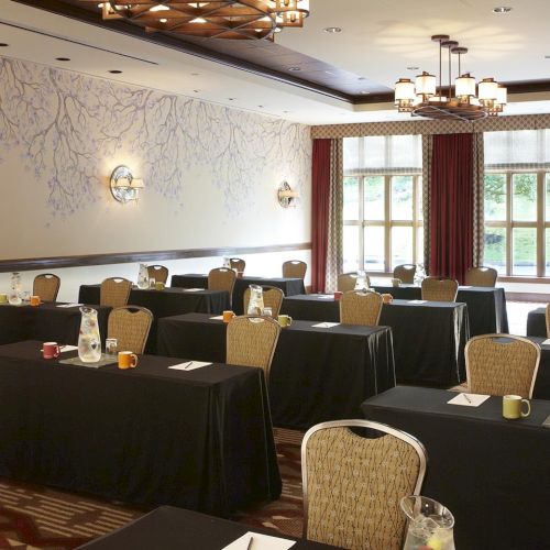 A bright conference room set with banquet tables, patterned chairs, black tablecloths, mugs, and chandeliers under large windows.