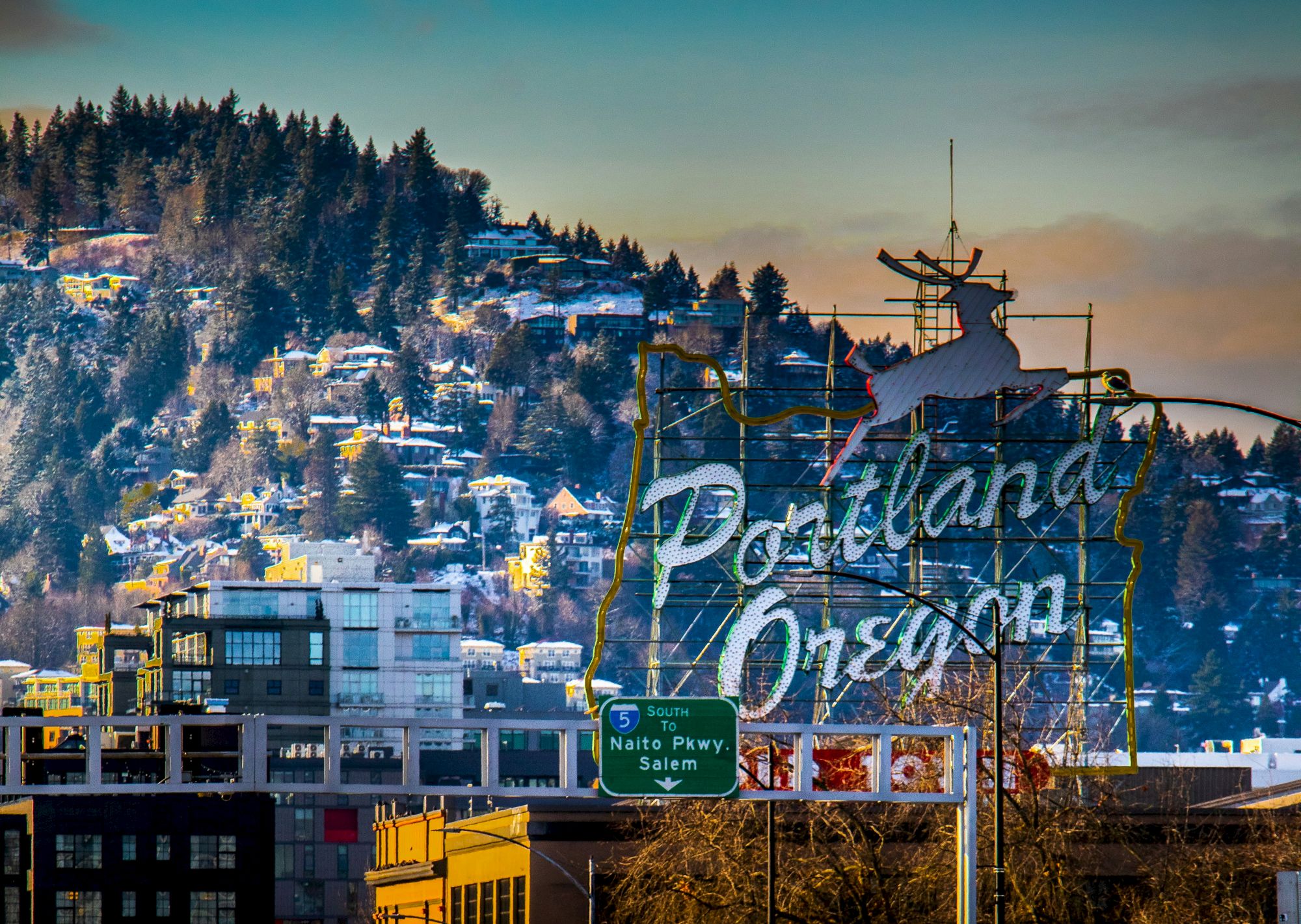 A winter city view with snowy hills and pines, and a large &ldquo;Portland&rdquo; sign on a bridge, with roadway directions below.