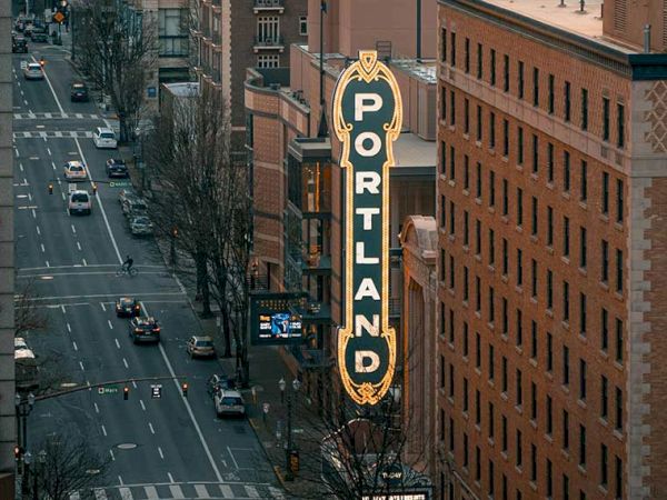 A city street between tall brick buildings, with a vertical &ldquo;PORTLAND&rdquo; sign hanging over the roadway.