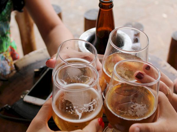 Friends clink amber beer glasses and toast together outdoors, with fizzy drinks and bubbles on a sunny patio.