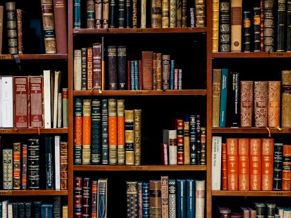 A wall of old, leather-bound and colorful books fills multiple wooden shelves in a library.