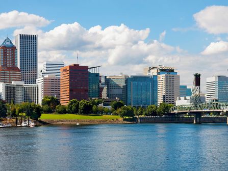 A waterfront city skyline with tall buildings, a bridge, and cranes over blue harbor waters under a partly cloudy sky.