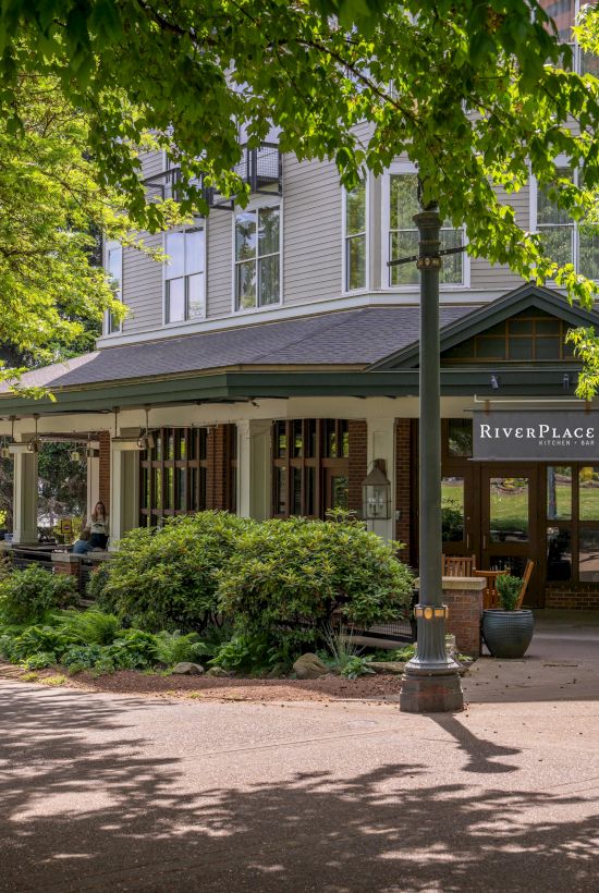 Sunny courtyard view of a brick-and-shingle building with the &ldquo;RiverPlace Kitchen Bar&rdquo; sign, framed by green trees.