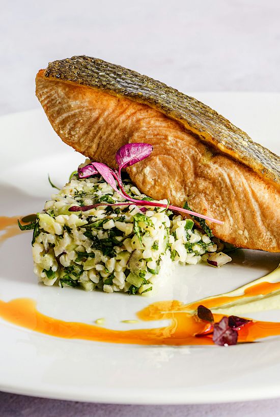 A plated grilled fish fillet with herb-and-rice salad, garnished with microgreens and drizzled orange and cream sauces.