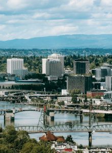 City skyline beside a wide river, with a large steel bridge and office towers under cloudy skies and distant hills.