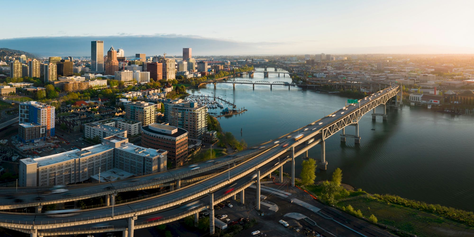 An aerial view of a modern city skyline with a wide river, bustling bridges, and golden-hour sunlight over downtown.