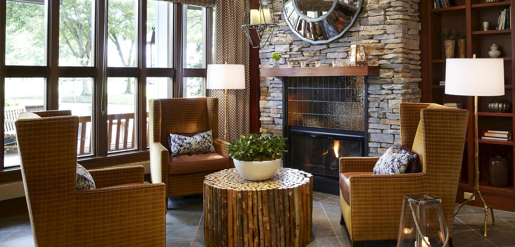 Cozy living room with a stone fireplace, two patterned armchairs, a wood-log coffee table, potted greenery, and big windows.