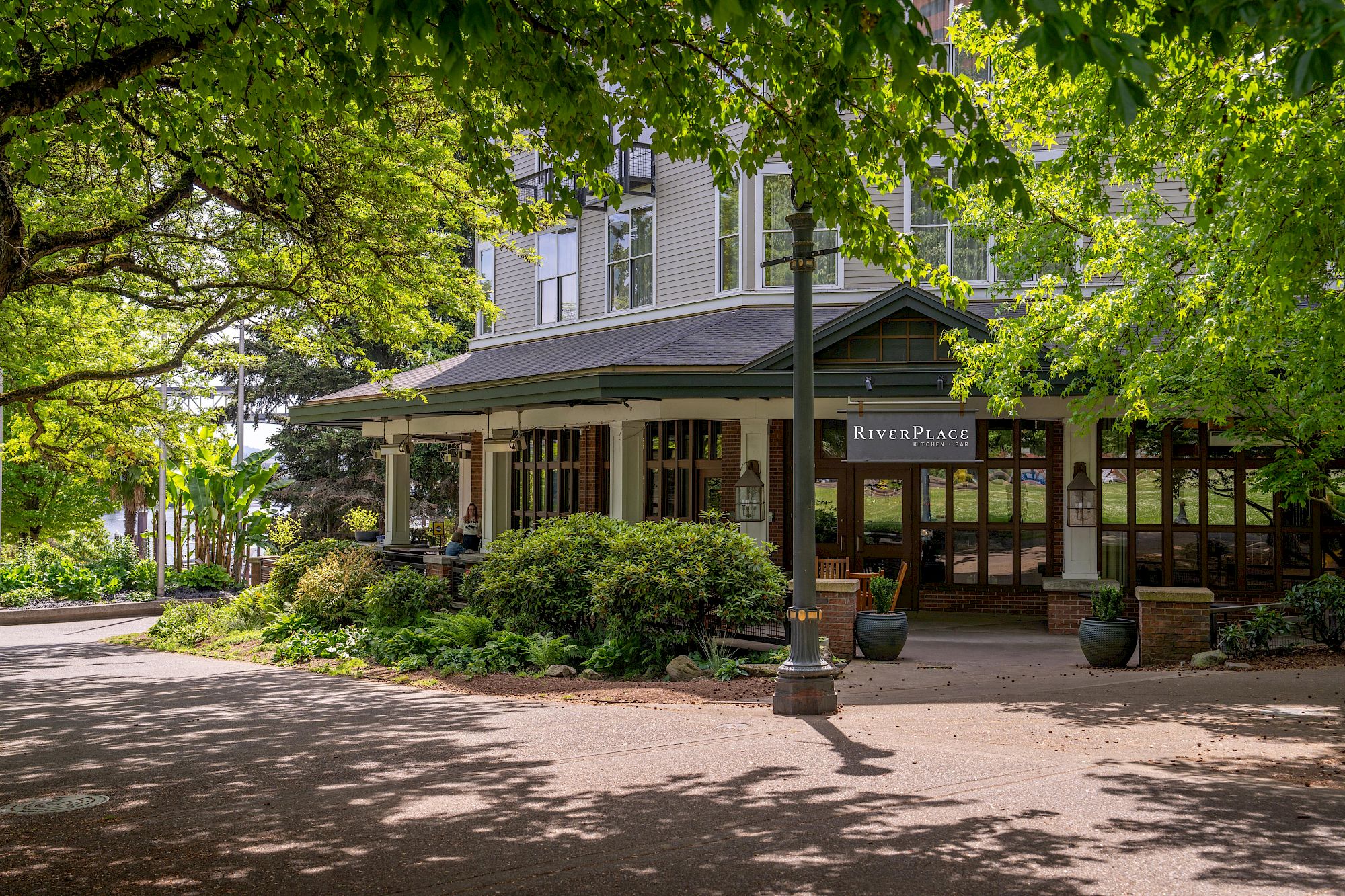 A sunny garden courtyard beside the &ldquo;RiverPlace Kitchen &bull; Bar,&rdquo; with leafy trees framing a brick-and-wood building.