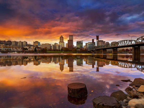 A city skyline at sunset with a calm river, reflections, and a steel bridge on the right.