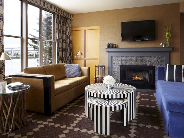 Cozy living room with blue seating, striped ottoman, and a lit fireplace beside large windows and patterned curtains.