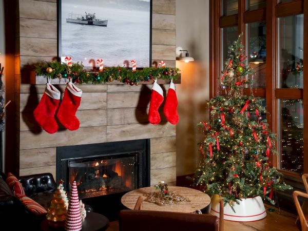 A cozy Christmas living room with a lit fireplace, wreath garland, red-and-white stockings, and a decorated tree by large windows.