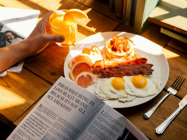 A breakfast plate with fried eggs, bacon, toast, and sliced fruit, plus orange juice being poured beside a newspaper.