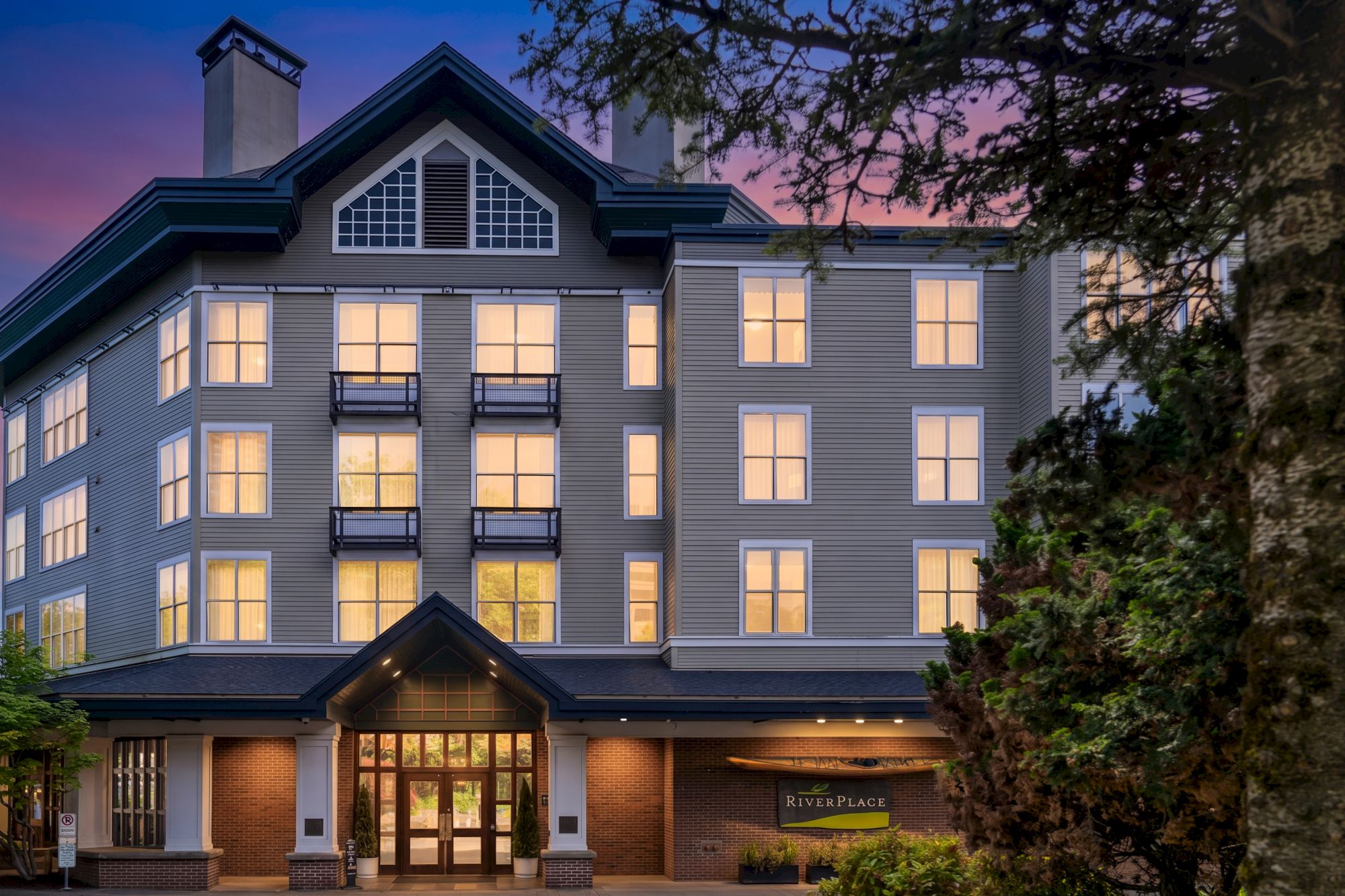 A modern apartment building with many windows and balconies at dusk, lit from within under a purple twilight sky.