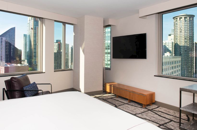 Modern hotel room with large windows, city skyline view, bed, TV, leather chair, and wooden media console.