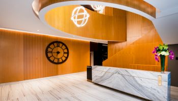 Modern hotel lobby with wood-paneled walls, marble reception desk, glowing globe light, wall clock, and a vase of flowers.