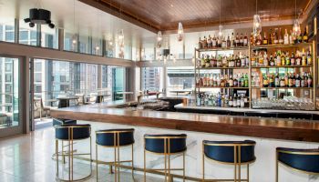 Modern bar interior with wooden ceiling, backlit shelves of liquor bottles, glassware, and marble counters with stools.