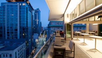 A modern rooftop terrace with lounge chairs overlooks tall city buildings and streets at dusk, showing a panoramic skyline view.