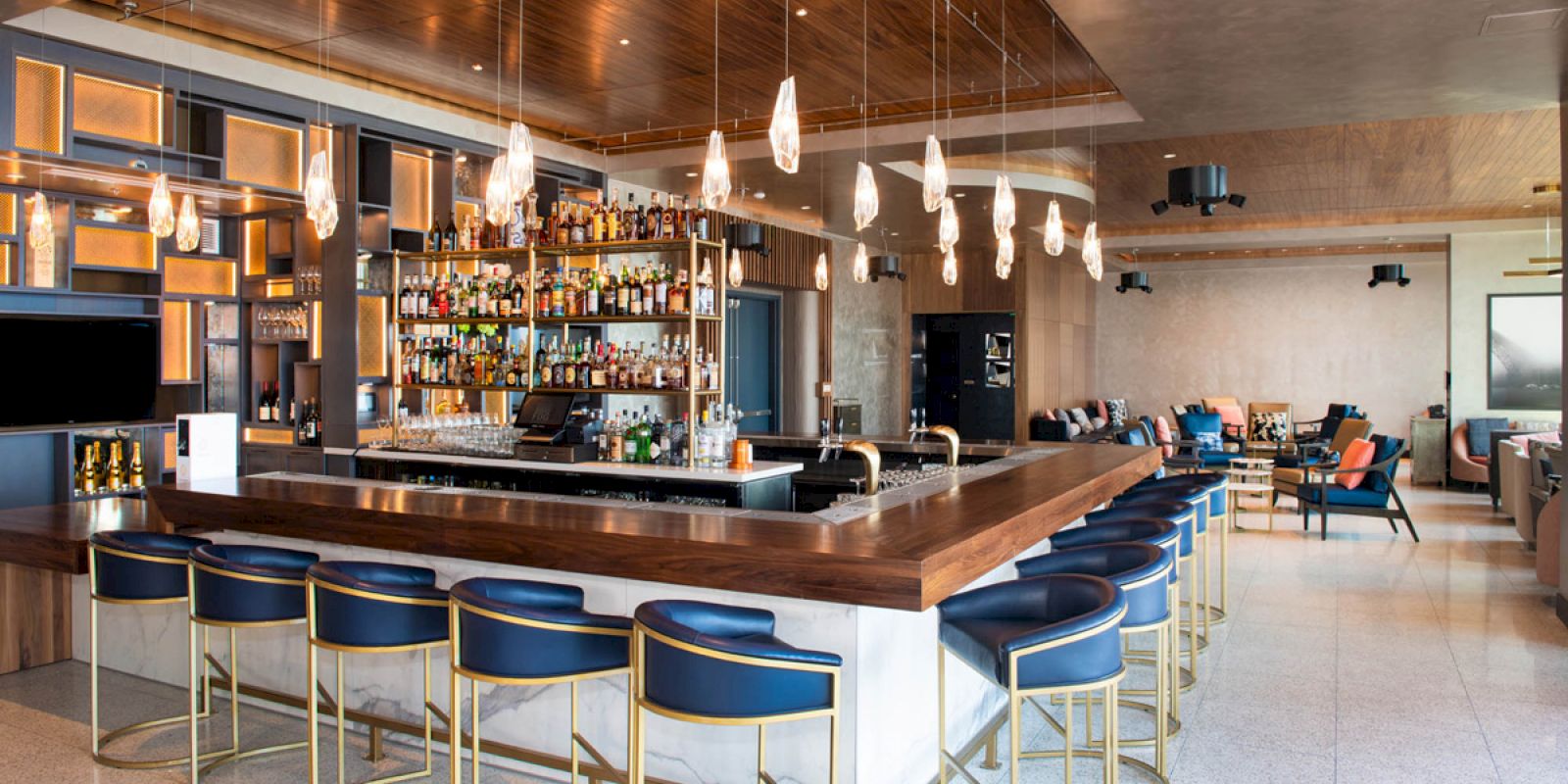 A modern bar with a wooden counter, backlit liquor bottles, blue stools, and hanging pendant lights in a lounge area.