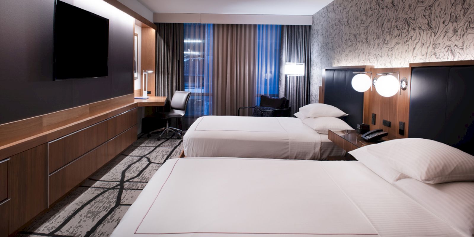 A modern hotel room with two beds, a wall-mounted TV, desk area, and warm lighting under blue curtains.