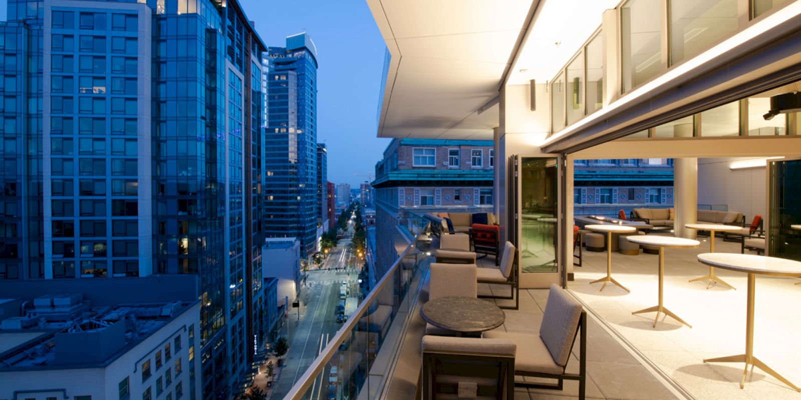 Balcony terrace with chairs and tables overlooking a downtown skyline of tall blue-lit buildings at dusk.