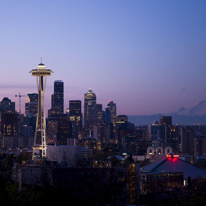 Seattle skyline at dusk with the Space Needle lit up, high-rises glowing against a pastel sky and distant mountains.