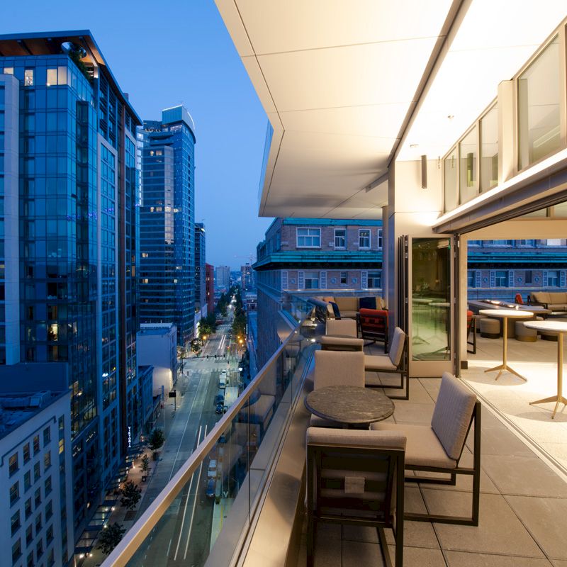 Rooftop terrace lounge with chairs and tables overlooking a downtown skyline of tall buildings under a twilight blue sky.