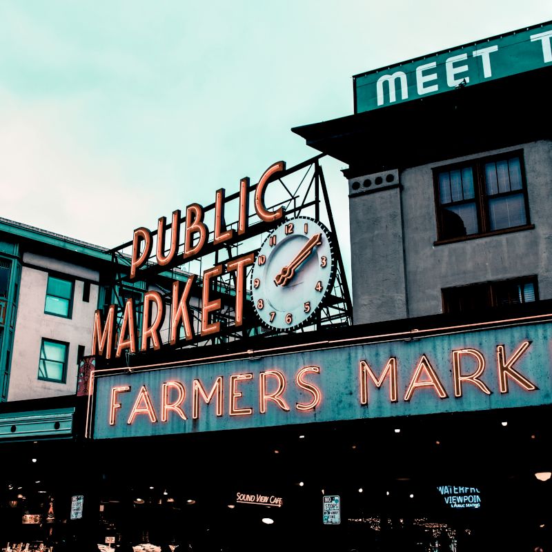 A neon &ldquo;Public Market&rdquo; clock sign on a building, with &ldquo;Farmers Market&rdquo; and &ldquo;Meet the Farmers&rdquo; text under the sky.