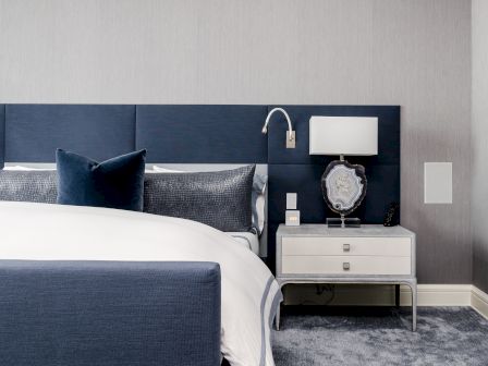 A modern hotel bedroom: blue upholstered headboard, pillows and bed, and a nightstand with lamp and decor.
