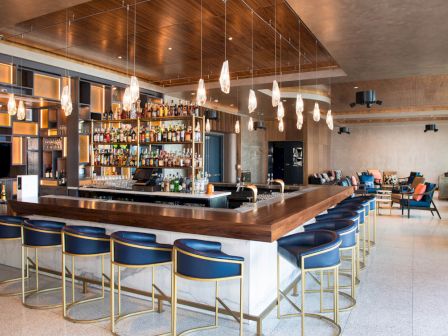 A modern bar lounge with a bottle-lined back bar, glowing pendant lights, and blue upholstered stools in a stylish seating area.