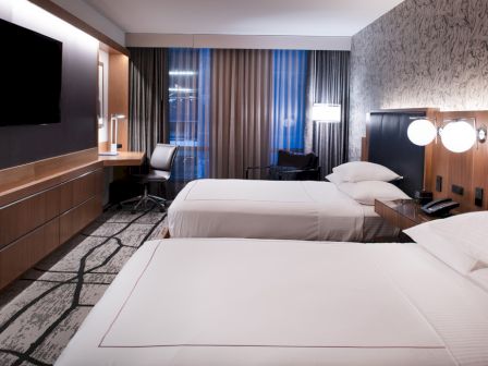 A modern hotel room with twin beds, a wall TV, dark curtains, and reading lights by the headboard.