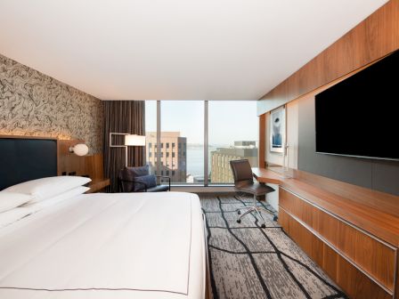 A modern hotel room with a bed, wall-mounted TV, wooden cabinetry, and large windows overlooking buildings.