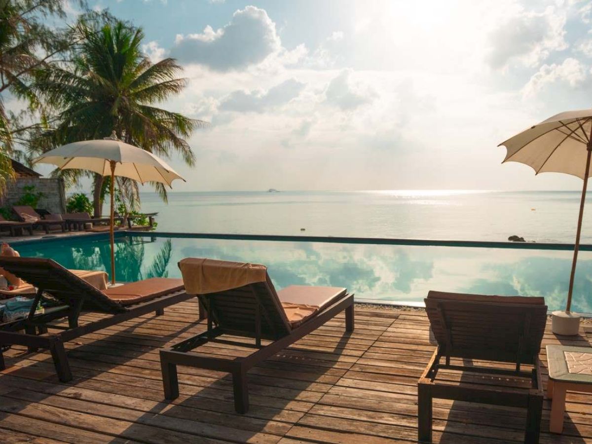 Beach resort scene with poolside lounge chairs, two umbrellas, palm trees, and calm ocean under a bright cloudy sky.