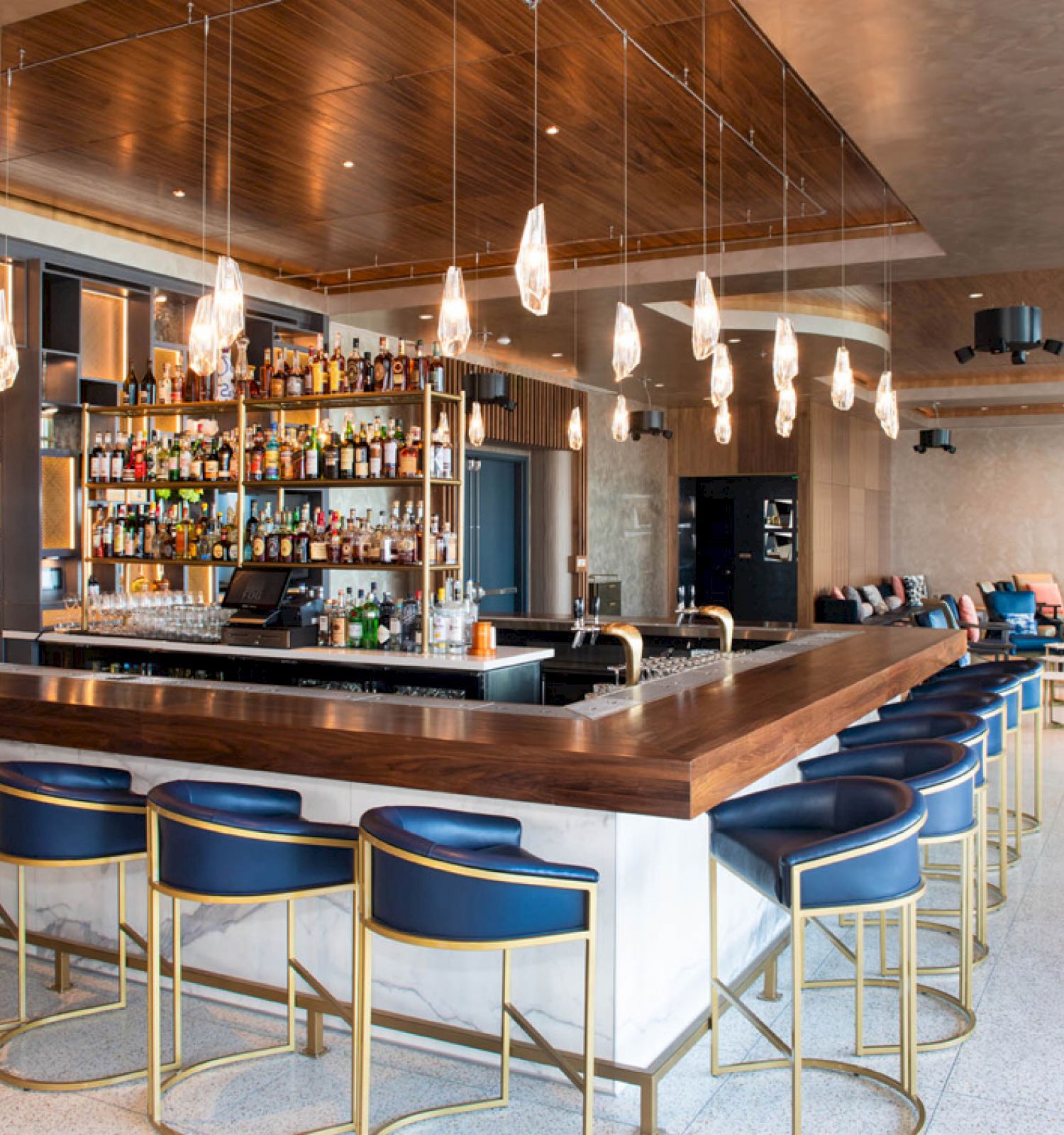 A modern hotel bar with backlit bottle shelves, glowing pendant lights, blue stools, and a lounge seating area.