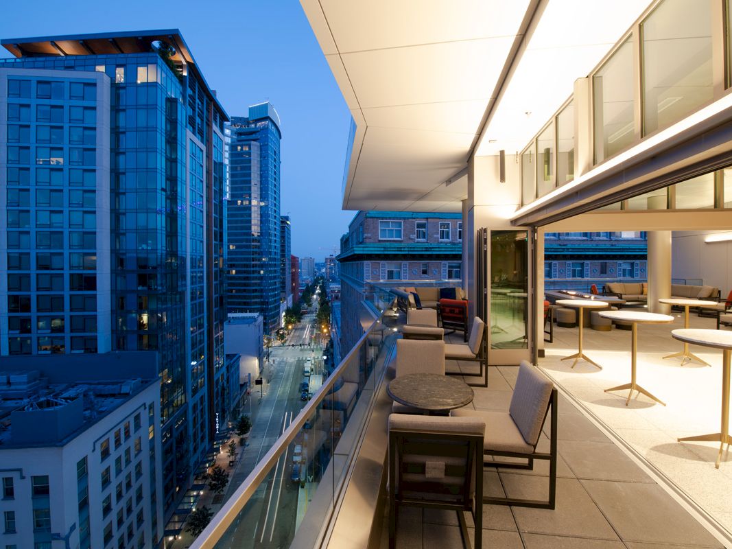 A modern rooftop terrace overlooks a city street at dusk, with lounge chairs and skyscrapers stretching into the distance.