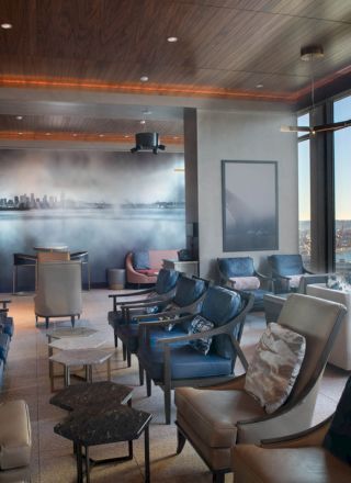 A modern lounge with leather chairs, low tables, and a floor-to-ceiling window overlooking a harbor at dusk.