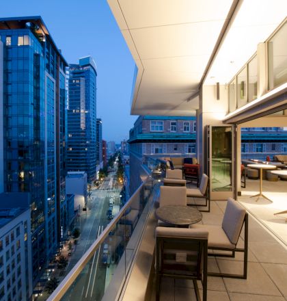Rooftop terrace with chairs and tables overlooks a downtown street lined by tall buildings at dusk.