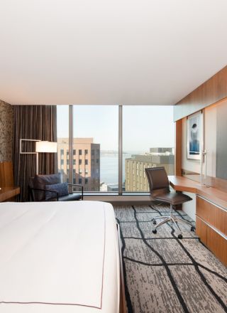 A modern hotel room with a large bed, wood cabinetry, a chair by the window, and a flat-screen TV.