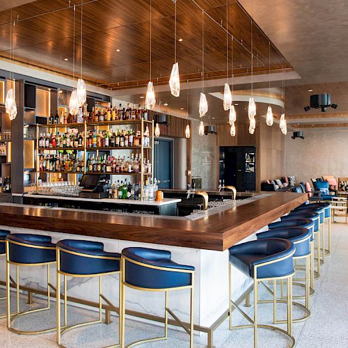 A modern hotel bar with a wooden counter, tall stools, hanging pendant lights, shelves of bottles, and lounge seating in the background.