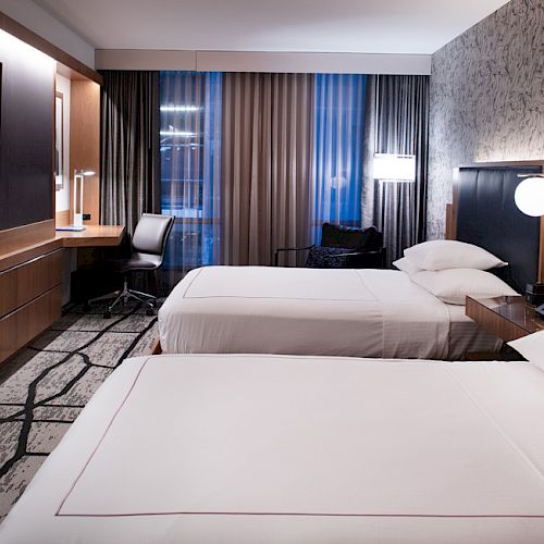 A modern hotel room with two neatly made beds, desk area, and large windows with curtains.