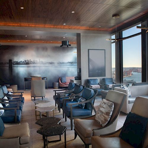 Modern lounge with plush chairs and couches, warm lighting, and large windows overlooking a river and city skyline.