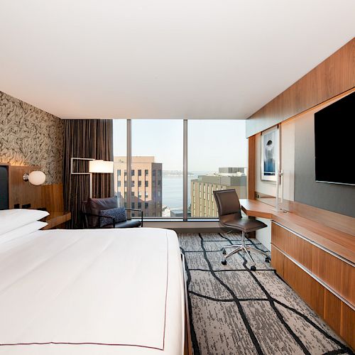 A modern hotel suite with a neatly made bed, a desk chair, wood cabinetry, flat-screen TVs, and large city-view windows.