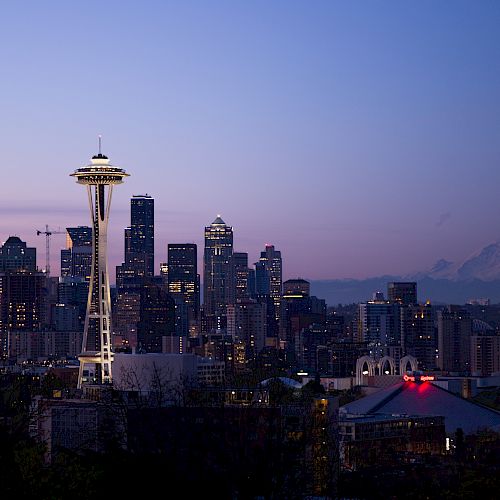 Seattle&rsquo;s skyline at dusk with the Space Needle and tall buildings lit up against a purple-blue sky.