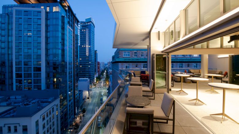 A modern rooftop terrace with outdoor seating overlooks a city of tall buildings at blue hour.