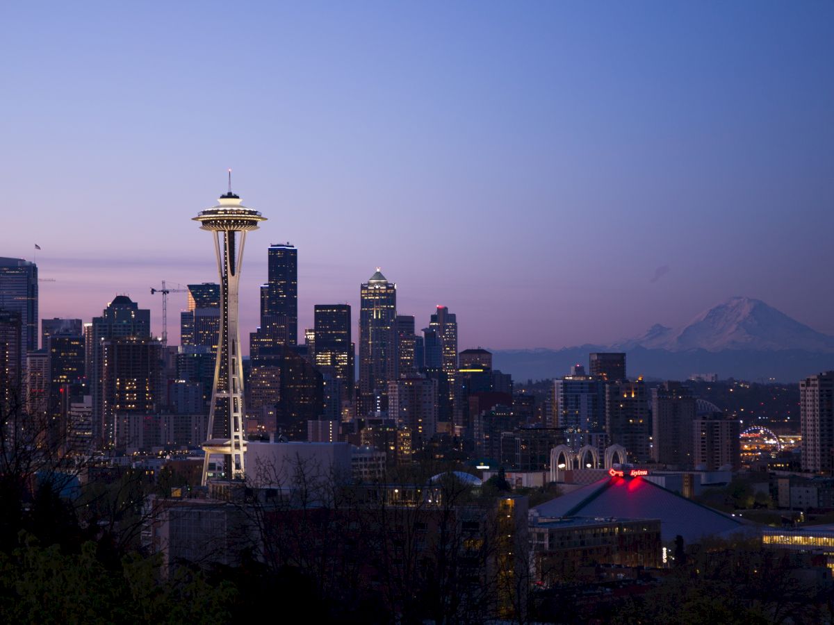 Seattle skyline at dusk with the Space Needle in the center, glowing city lights and distant mountains under a purple sky.