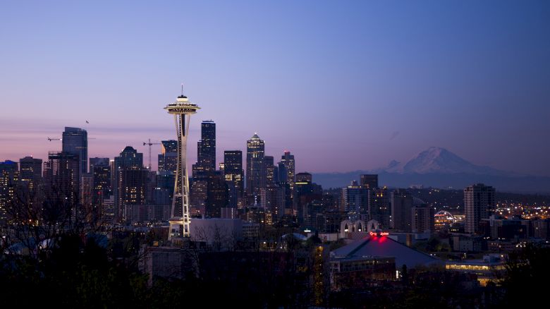 Seattle skyline at dusk with the Space Needle in the center, glowing city lights and distant mountains under a purple sky.