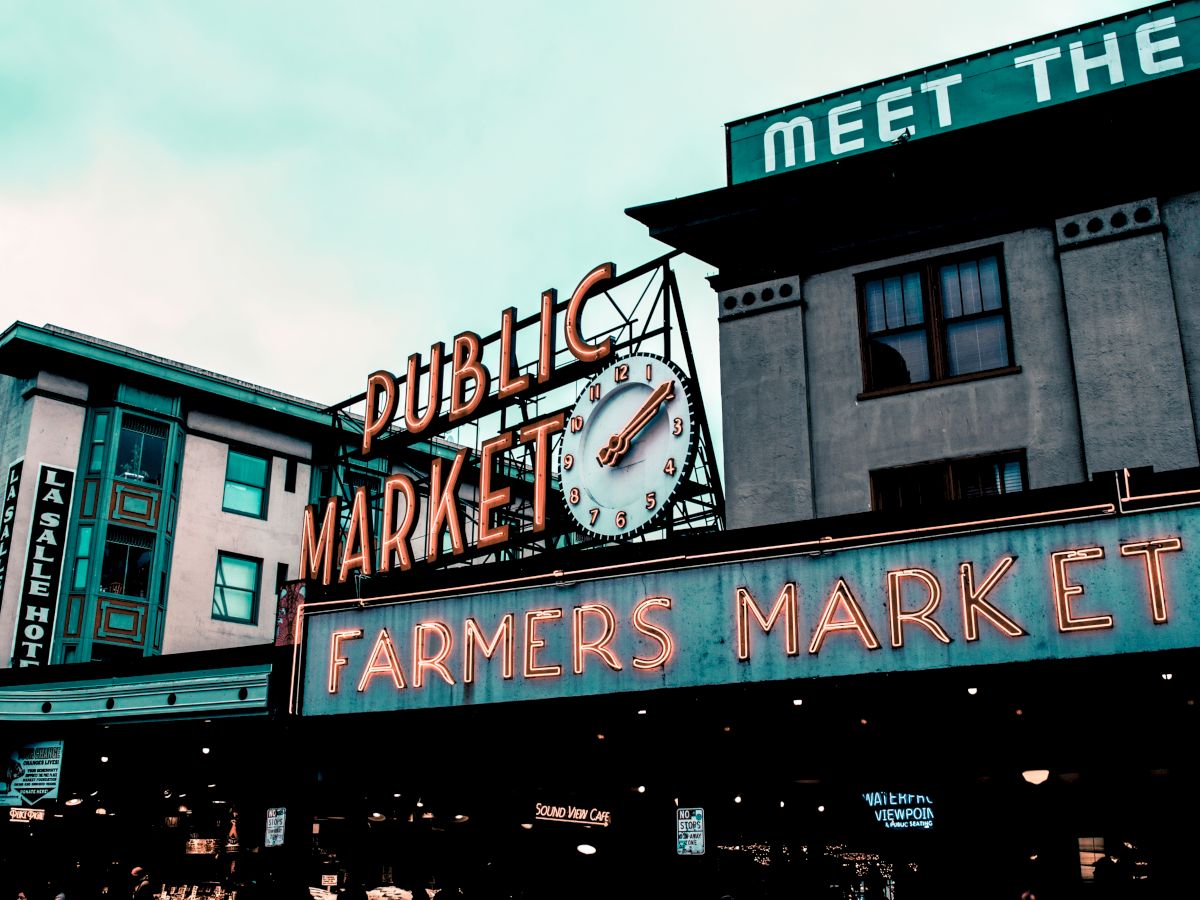 A neon-lit clock and sign reading &ldquo;DUBLINIC&rsquo;S MARKET,&rdquo; with &ldquo;FARMERS MARKET&rdquo; and storefronts under a bright sky.