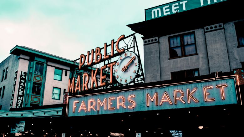A neon-lit clock and sign reading &ldquo;DUBLINIC&rsquo;S MARKET,&rdquo; with &ldquo;FARMERS MARKET&rdquo; and storefronts under a bright sky.