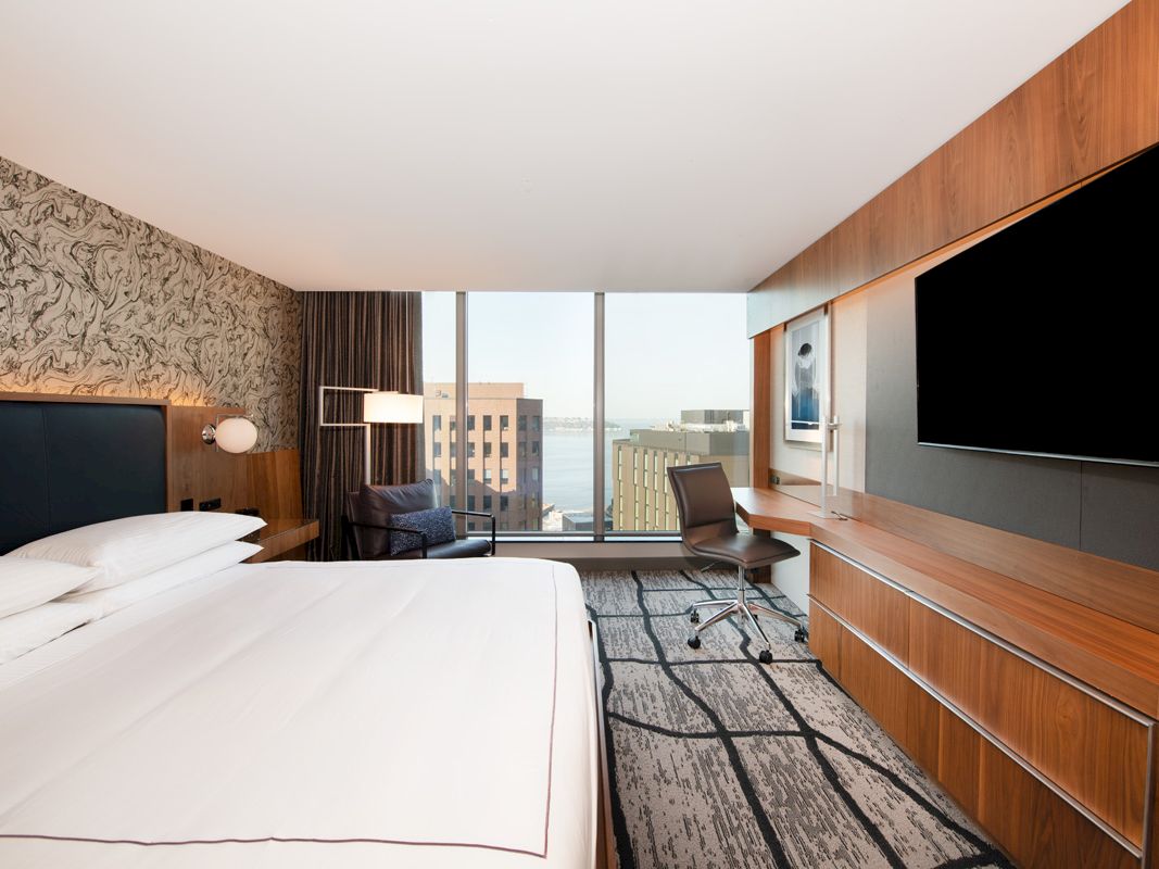 A modern hotel room with a large bed, wood wall panels, desk chair, TV, and big windows overlooking buildings.