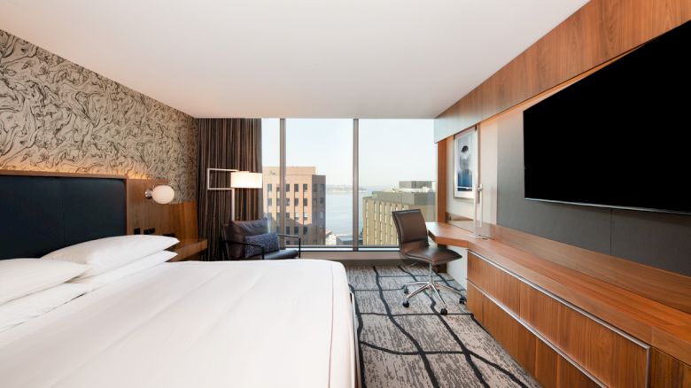A modern hotel room with a large bed, wood wall panels, desk chair, TV, and big windows overlooking buildings.